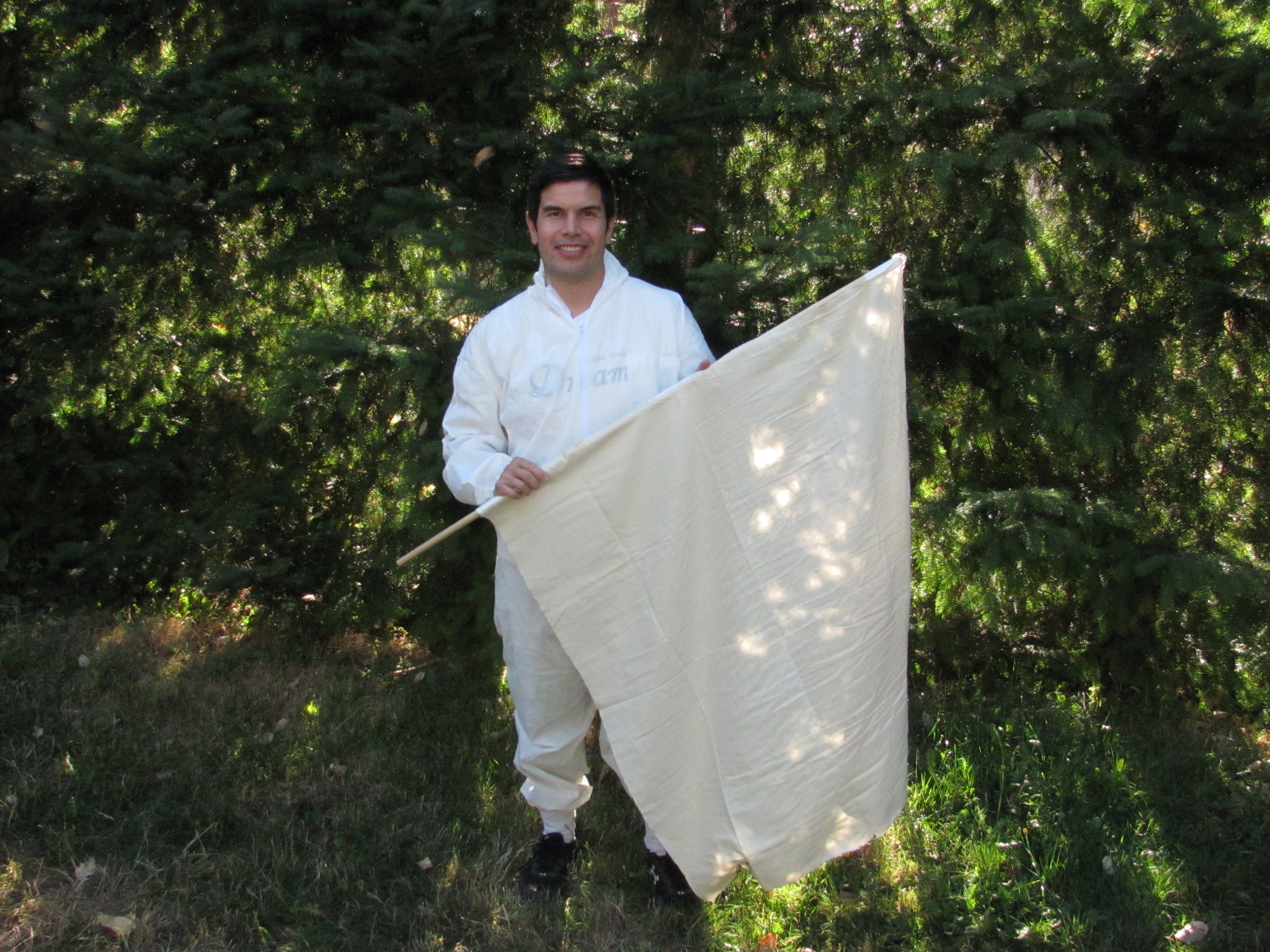 A man stands in a white flannel suit in the forest, holding a white blanket used to collect ticks.