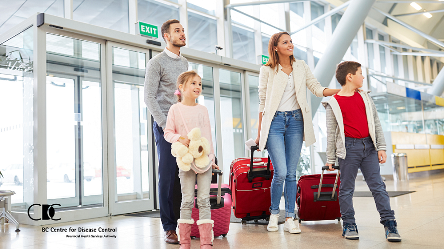 A family of two adults and two young children stand smiling in an airport while carrying rolling suitcases.