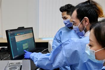 Three lab technicians wearing gowns and surgical masks look at a screen in a lab