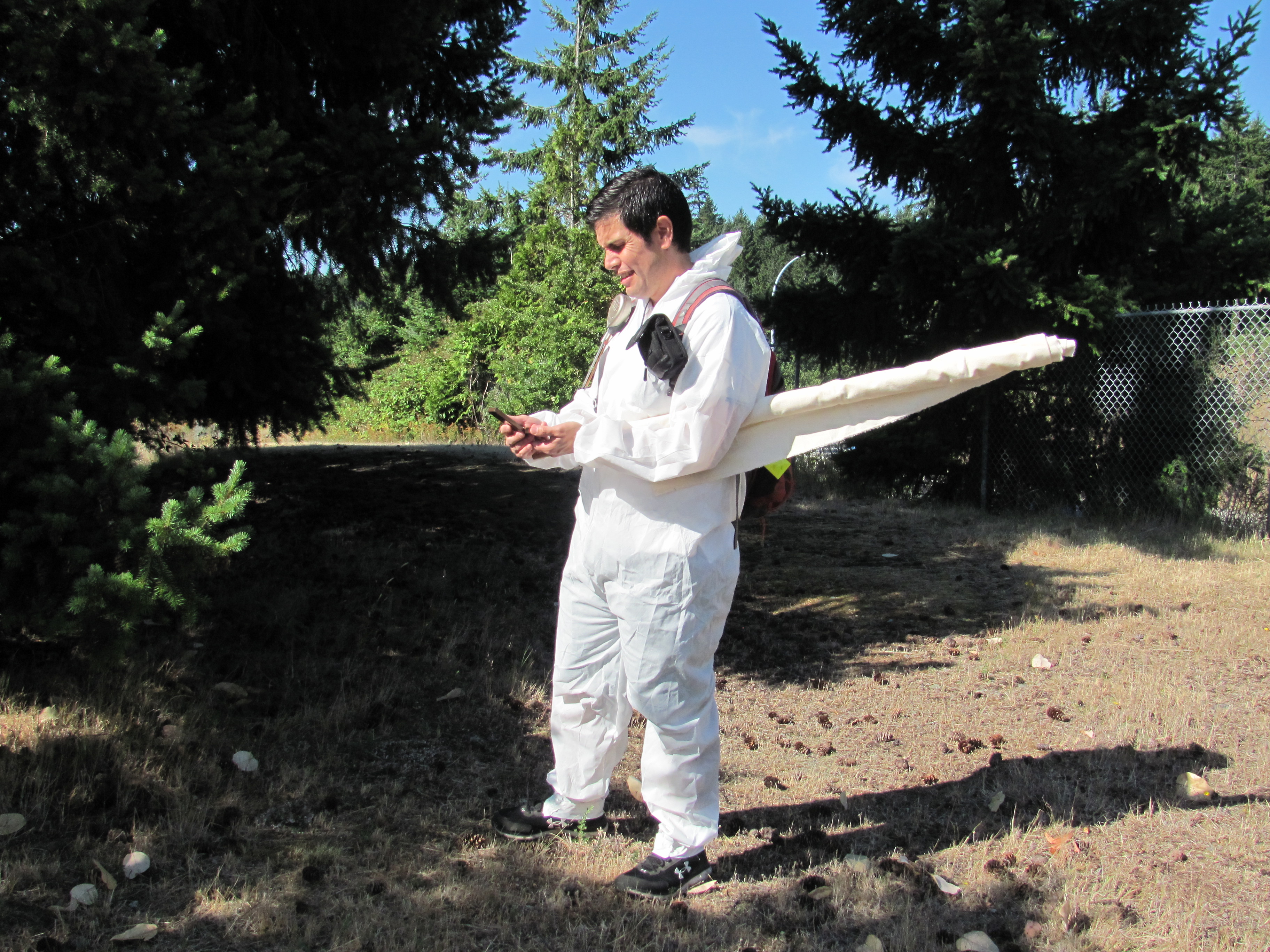 A man stands in a white flannel suit in the forest, with a white blanket tucked under his arm.