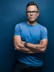 A man with styled black hair, glasses and a light blue t-shirt stands and smiles