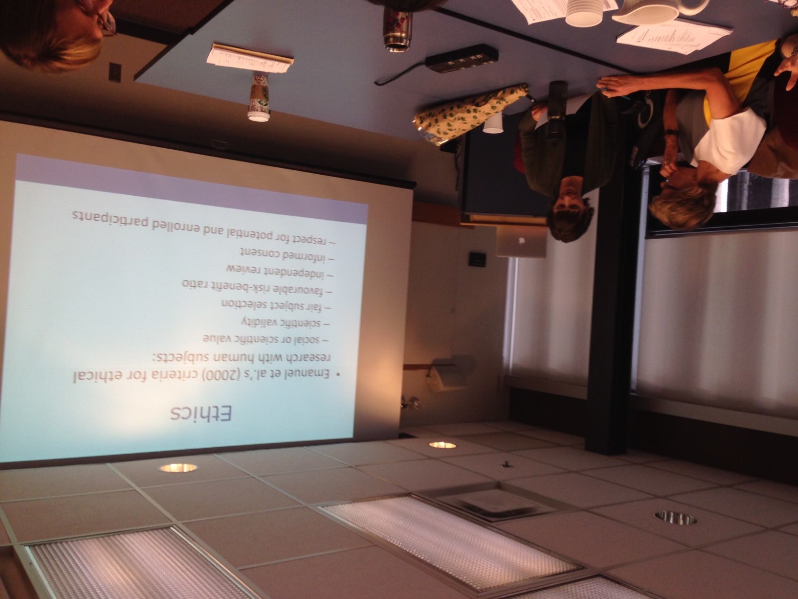 Two women are seated at a table looking at a piece of paper, a powerpoint screen reads "Ethics"